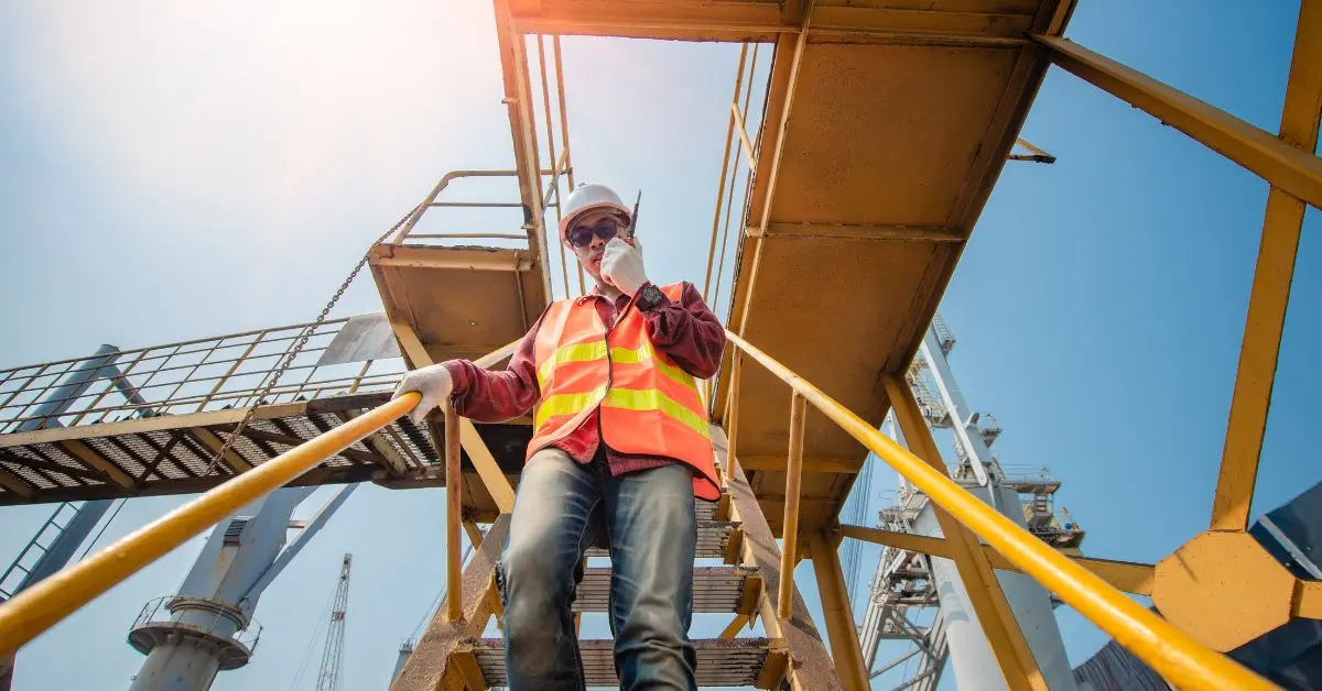 Un trabajador con casco y chaleco reflectante de seguridad subiendo una escalera metálica en una zona industrial, comunicándose a través de una radio.